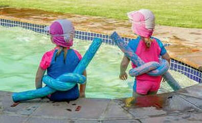 Picture of the back of 2 children sitting on the side of the pool with their legs in the pool, they are wearing bandanas and goggles with twist floaties around thier waists